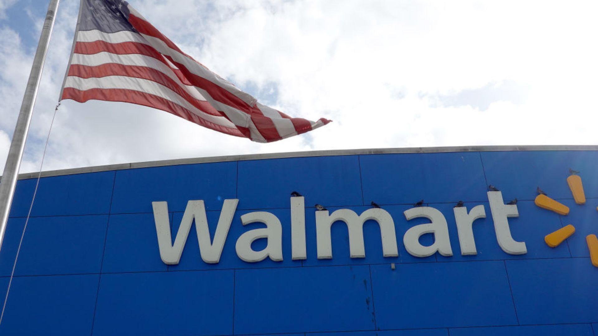 A Walmart storefront with the store's name in large white and yellow letters against a blue background. Above the store, an American flag is waving on a flagpole