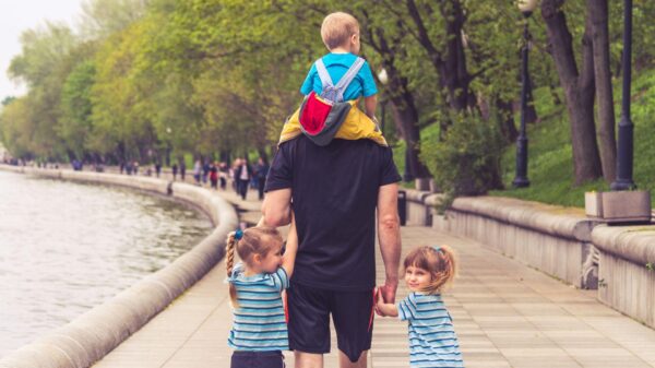 A father walks along a riverside pathway with his young son sitting on his shoulders and holding hands with his two daughters