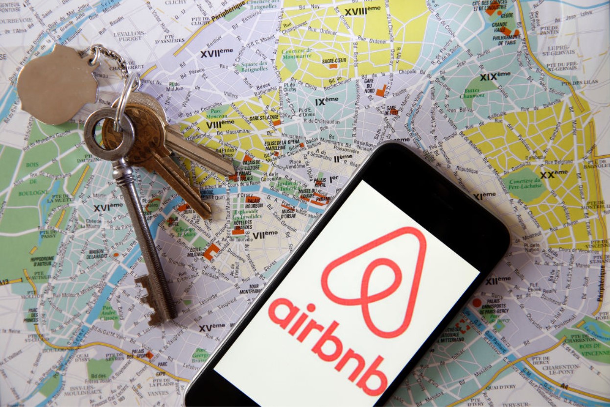 Airbnb logo is displayed on the screen of an iPhone placed on a map of the city of Paris with a set of keys