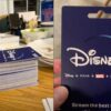 A photograph displaying a person's hand holding a stack of blue Disney+ gift cards, each valued at $100, with the logo and tagline "Stream the best stories" visible on one card held at the forefront. On the right hand side of the image is a close-up view of the gift card