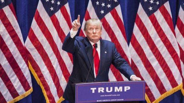 Donald Trump in a blue suit with a red tie pointing as he stands in front of three American flags.