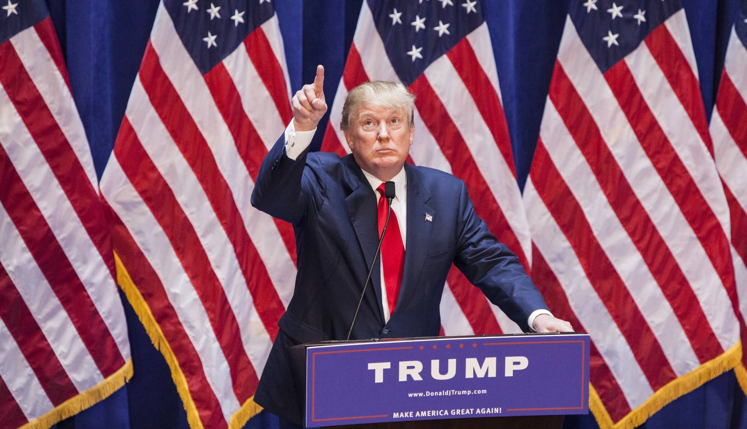 Donald Trump in a blue suit with a red tie pointing as he stands in front of three American flags.